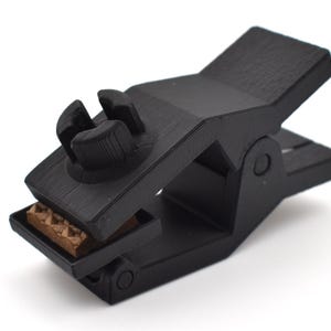 Pode incluir: Um grampo de mola preto com acabamento fosco texturizado, um botão de ajuste central com topo de quatro pontas e uma pequena inserção retangular marrom posicionada na mandíbula inferior do mecanismo para uma fixação segura.