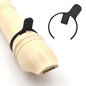 Può includere: Primo piano di un flauto dolce beige con un poggia-pollice in plastica nera attaccato. È visibile anche un poggia-pollice in plastica nera separato. Sfondo bianco.