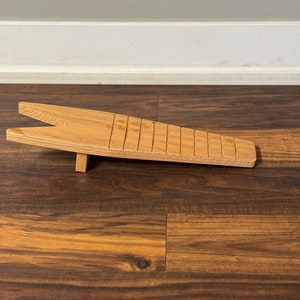 May include: A wooden guitar fretboard notched with evenly spaced slots. The fretboard is light brown and rests on a small wooden support. The background is a wooden floor and a white wall.