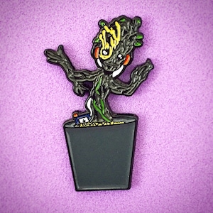 以下が含まれることがあります： ガーディアンズ・オブ・ギャラクシー・フランチャイズの架空のキャラクター、グルートを描いたエナメルピン。グルートは、頭に鉢を乗せたヒューマノイドの木として描かれています。このピンは、マーベル・シネマティック・ユニバースのファン向けの収集品です。