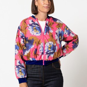 Könnte beinhalten: Eine pinkfarbene Bomberjacke mit floralem Muster, blauem Samtbesatz und Reißverschluss. Die Jacke hat ein lebendiges Muster aus blauen, weißen und goldenen Blumen. Das Model trägt eine dunkelblaue Jeans.