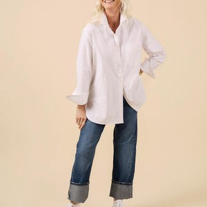 May include: A woman wearing a white button-down shirt and blue jeans with cuffed hems. She is also wearing white high-top sneakers. The shirt has long sleeves and a classic collar. The jeans are a medium wash denim.