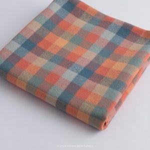 Puede incluir: Tela doblada con un estampado de cuadros en tonos naranja, azul y beige. La tela es cuadrada y parece estar hecha de un material suave. El patrón es un diseño de cuadros clásico.