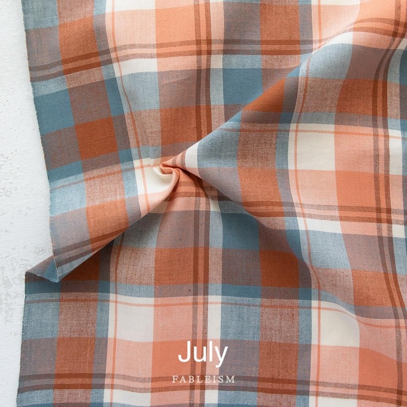 K&ouml;nnte beinhalten: Nahaufnahme eines Stoffes mit Karomuster in Orange-, Blau-, Wei&szlig;- und Braunt&ouml;nen. Das Wort "July" ist wei&szlig; gedruckt, darunter "FABLEISM". Der Stoff ist gefaltet und drapiert.