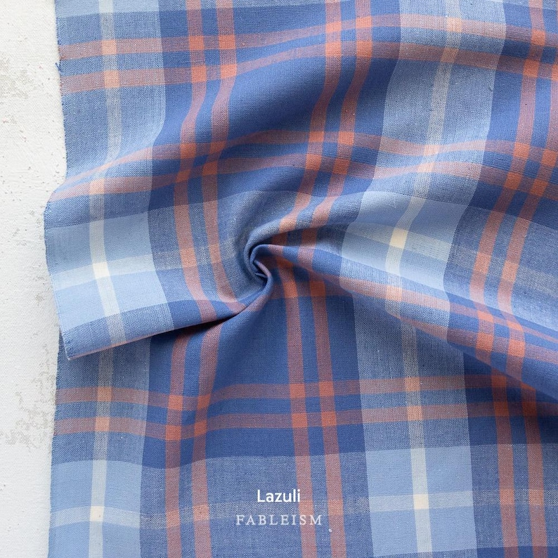 a close-up view of a fabric with a plaid pattern in shades of blue, orange, and white. The fabric appears to be folded or crumpled, creating a textured and visually interesting surface.