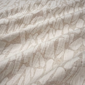 Könnte beinhalten: Nahaufnahme eines beige-weißen Stoffes mit strukturiertem Muster. Der Stoff hat ein gewebtes Aussehen mit einem sich wiederholenden Design aus weißen, unregelmäßigen Formen auf beigem Hintergrund. Der Stoff ist drapiert und zeigt Falten und Schatten.