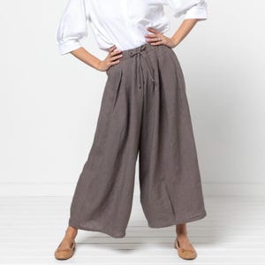 May include: Wide-leg, brown linen pants with a drawstring waist. The pants are paired with a white button-down shirt and tan flats. The pants have pleats at the waist and a relaxed fit.
