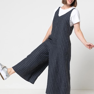 May include: A woman wearing a navy blue and white striped jumpsuit with a white t-shirt underneath. She is standing with one leg raised and is wearing silver sneakers.