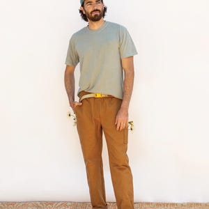 May include: A brown pair of pants with a yellow belt and a light blue t-shirt. The pants are called "The Rambler Pants" and are from the company "FRIDAY pattern co.".