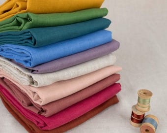 May include: A stack of folded fabric squares in various colors including mustard yellow, forest green, teal, blue, lavender, beige, light pink, magenta, and rust, accompanied by three small wooden spools wrapped with colored sewing thread.