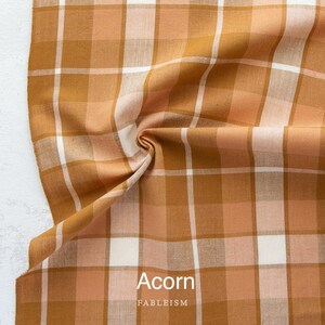 K&ouml;nnte beinhalten: Nahaufnahme eines Stoffes mit Karomuster in Braun-, Pfirsich- und Wei&szlig;t&ouml;nen. Der Stoff ist gefaltet und zeigt die Textur und Anordnung der Farben. Das Wort "Acorn" ist auf den Stoff gedruckt, darunter der Markenname "FABLEISM".