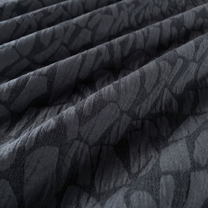 May include: Close-up of a dark gray fabric with a textured, geometric pattern. The fabric is folded and has a soft, wrinkled appearance.