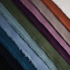 May include: A close-up of a variety of corduroy fabrics in different colors, including black, navy blue, teal, light blue, green, brown, purple, pink, gray, and white. The fabrics are arranged in a diagonal pattern, with the edges of each fabric piece frayed.