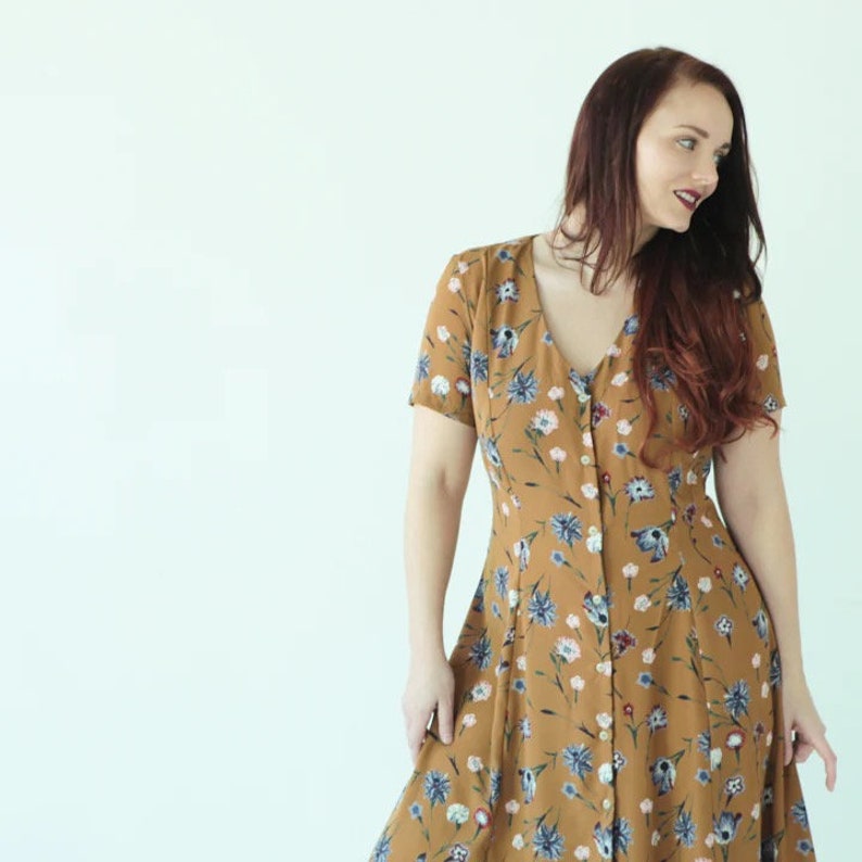 May include: A woman wearing a brown floral dress with a v-neck and short sleeves. The dress has a button-up front and a fitted waist.