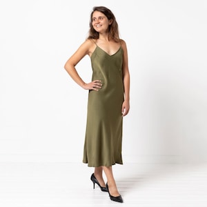May include: Olive green slip dress with spaghetti straps and a V-neck. The midi-length dress has a slight A-line shape and is paired with black high heels. The model is smiling and posing in front of a white background.