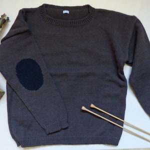 Peut inclure: Un pull en tricot marron avec un coude noir. Le pull est posé à plat sur une surface blanche avec deux aiguilles à tricoter en bois.