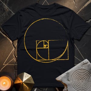Könnte beinhalten: Ein schwarzes T-Shirt mit einer goldenen Umrandung der Fibonacci-Spirale, einem mathematischen Konzept, das Harmonie und Gleichgewicht darstellt.