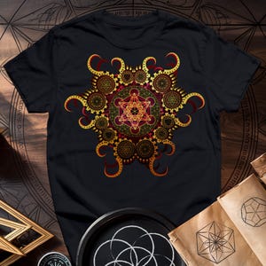 May include: Black t-shirt featuring a gold and red geometric design. The design incorporates a star within a circle, surrounded by smaller circles and intricate patterns.