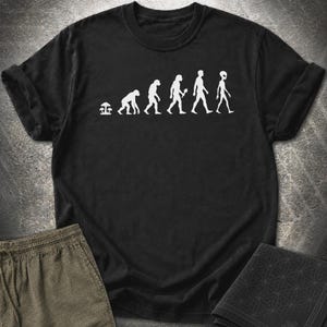 May include: A black short-sleeved t-shirt featuring a white graphic print that depicts the evolutionary progression starting with mushrooms, followed by primates, early humans, and ending with a tall, slender alien figure in a walking sequence.