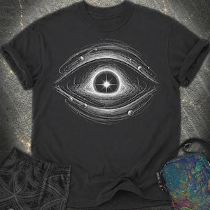 Può includere: Una t-shirt nera a maniche corte con una stampa grafica bianca che raffigura una forma d'occhio stilizzata composta da anelli cosmici vorticosi e piccole sfere planetarie, con un punto di luce brillante al centro della pupilla.