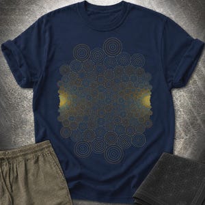 Può includere: Una t-shirt a maniche corte blu navy con un design grafico centrale composto da numerosi cerchi concentrici nelle tonalità oro, bronzo e azzurro. Il motivo circolare è denso, creando un effetto geometrico che si irradia dal centro verso i lati.
