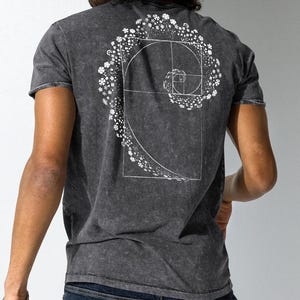 Peut inclure: Un t-shirt gris foncé avec un motif blanc en forme de spirale composé de fleurs. La spirale est à l'intérieur d'un carré.