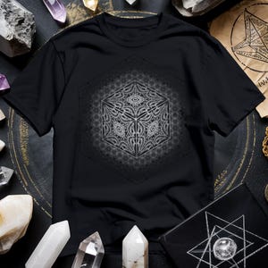 May include: Black t-shirt featuring a detailed white geometric cube design within a hexagon, surrounded by a circular pattern. The intricate design is centered on the shirt, creating a striking visual contrast against the dark fabric.