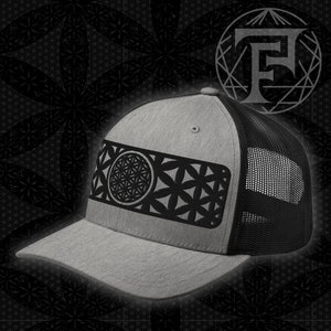 May include: A gray and black trucker hat with a black geometric design on the front. The design features a circle with a flower-like pattern inside.