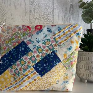 May include: A patchwork fabric zippered pouch with a white tassel. The pouch is made of various floral and geometric patterned fabrics in shades of blue, yellow, and white.