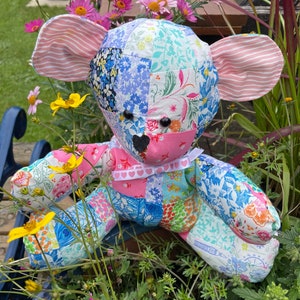 May include: A patchwork fabric teddy bear with pink ears and a pink bow with white hearts. The bear is sitting in a pot of pink and yellow flowers.