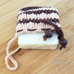 May include: A crocheted soap saver with brown and beige stripes and brown tassels. The soap saver is holding a bar of white soap. The soap saver has brown beads on the tassels. The soap saver is on a wooden surface. Stuart Soap Co.
