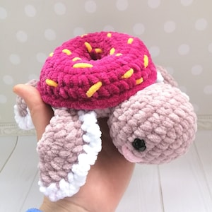 Peut inclure: Un jouet en crochet en forme de tortue en forme de beignet rose et jaune. La tortue a une carapace rose avec des pépites jaunes et un corps rose clair avec des accents blancs.