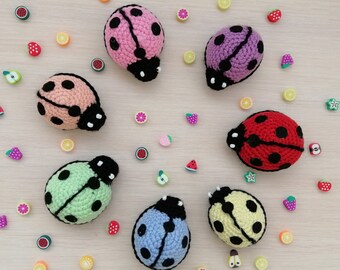 May include: A collection of seven crocheted ladybirds in various colours, including pink, purple, green, yellow, red, and blue. The ladybirds are surrounded by small decorative items, such as strawberries, watermelons, and lemons.