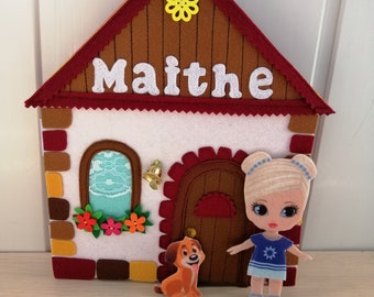 Könnte beinhalten: Ein Filzhaus mit einem roten Dach, braunen Wänden und einer weißen Tür. Das Haus hat ein Fenster mit einem blau-weißen Vorhang und eine gelbe Blume auf dem Dach. Der Name "Maithé" steht auf der Vorderseite des Hauses. Eine Mädchenpuppe mit blonden Haaren und einem blauen Kleid steht neben dem Haus, und ein brauner Hund steht vor dem Haus.