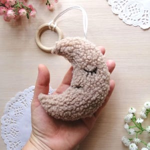 Pode incluir: Uma lua crescente de pel&uacute;cia bege com olhos bordados, presa a um anel de madeira com um cord&atilde;o branco.