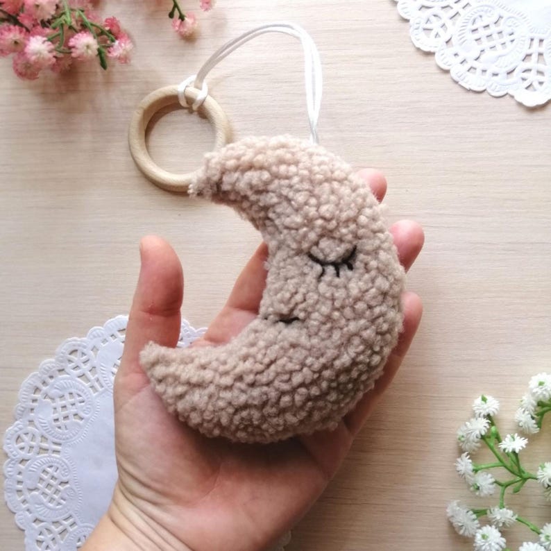 Pode incluir: Uma lua crescente de pel&uacute;cia bege com olhos bordados, presa a um anel de madeira com um cord&atilde;o branco.