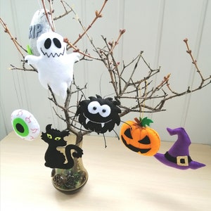 May include: Five felt Halloween ornaments hanging from a branch. The ornaments are a ghost, a spider, a pumpkin, a black cat, and a witch's hat. The ornaments are all handmade and have a whimsical, festive feel.