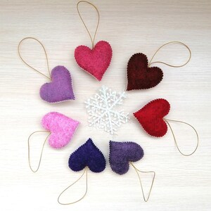 May include: Seven felt heart ornaments in various shades of pink, purple, and red, with gold string attached to each. The hearts are arranged in a circle on a light wood background.