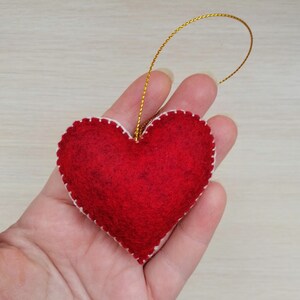 May include: A red felt heart ornament with white stitching around the edges. The heart is attached to a gold string.