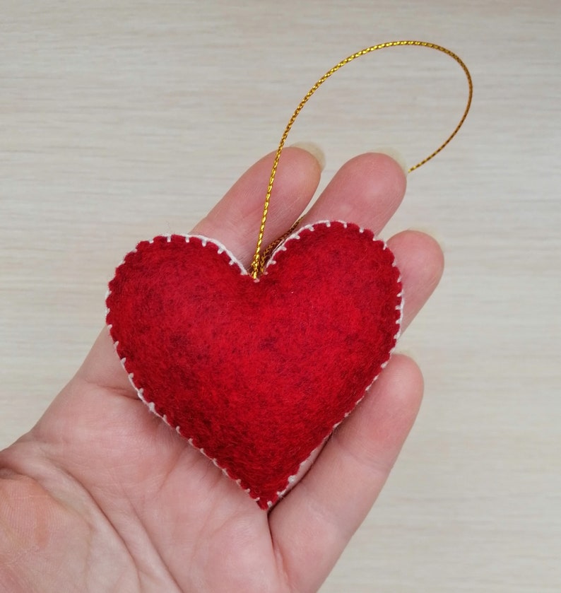 May include: A red felt heart ornament with white stitching around the edges. The heart is attached to a gold string.