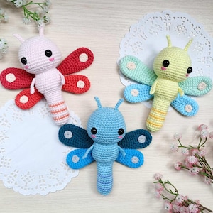Puede incluir: Tres juguetes de mariposa de ganchillo hechos a mano en rosa, azul y amarillo. Cada mariposa tiene alas grandes y coloridas y una cara sonriente con ojos negros. Los juguetes se exhiben sobre una superficie blanca con detalles florales.