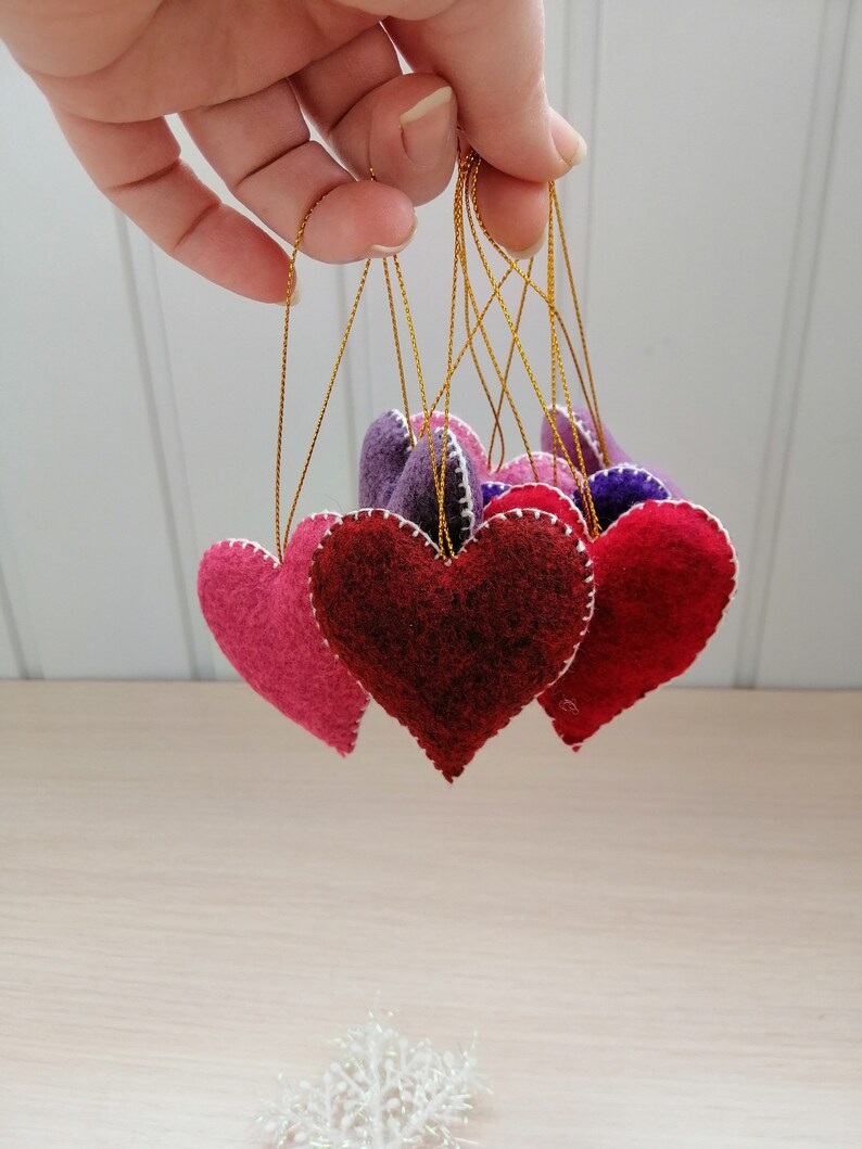 May include: Five felt heart ornaments in shades of pink, red, and purple, hanging from gold strings.