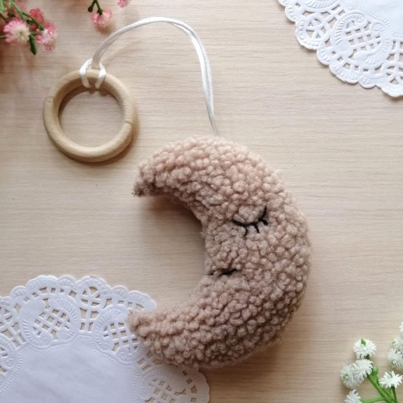 Pode incluir: Uma lua crescente de pel&uacute;cia bege com olhos bordados, presa a um anel de madeira com um cord&atilde;o branco.