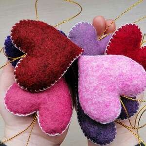 May include: A collection of felt heart ornaments in various shades of pink, purple, and red. The hearts are stitched with white thread and have a gold string attached for hanging.