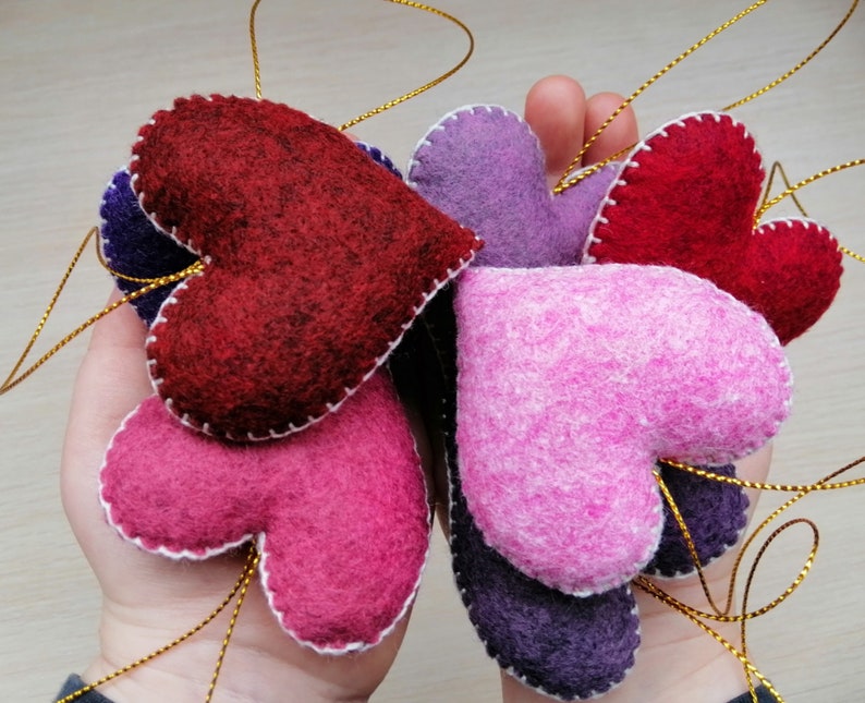 May include: A collection of felt heart ornaments in various shades of pink, purple, and red. The hearts are stitched with white thread and have a gold string attached for hanging.