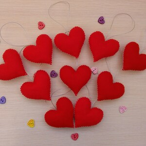 May include: Nine red felt hearts with white stitching, each hanging from a silver string. The hearts are arranged in a circle on a light brown wooden surface. There are small colorful heart-shaped buttons scattered around the hearts.