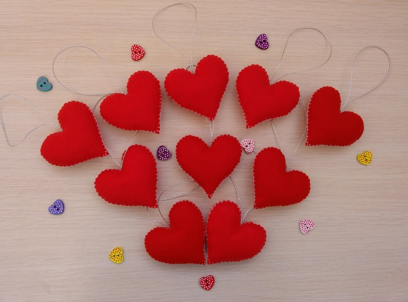 May include: Nine red felt hearts with white stitching, each hanging from a silver string. The hearts are arranged in a circle on a light brown wooden surface. There are small colorful heart-shaped buttons scattered around the hearts.