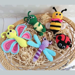 May include: Five crocheted stuffed animals in a wicker basket. The animals are a butterfly with pink and blue wings, a green caterpillar, a yellow and black bee, a purple and blue dragonfly, and a red ladybug.