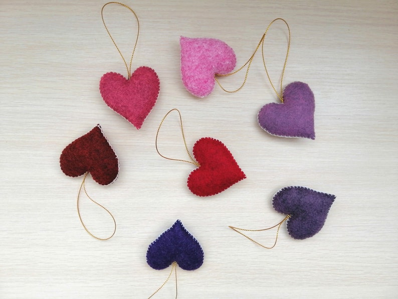 May include: Seven felt heart ornaments in various colors, including red, pink, purple, and burgundy. Each heart has a gold string attached for hanging.