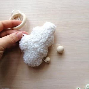 Pode incluir: Brinquedo de pel&uacute;cia branco e felpudo em forma de nuvem com contas de madeira penduradas. O brinquedo est&aacute; preso a um anel de madeira.